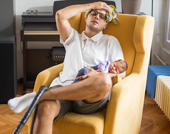 Dad with headache and tired, is asleep in chair with baby in his arm. This is not a SafeSleep360° recommended sleep position.